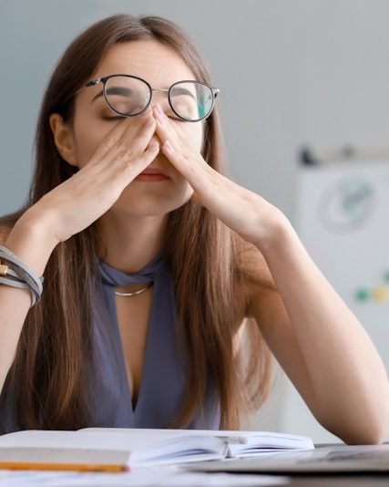 Eine erschöpfte Frau sitzt am Schreibtisch und reibt sich die Augen